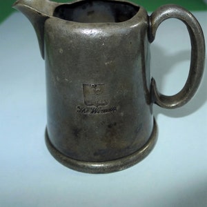 May include: A small, antique silver-coloured jug with a spout and handle. The jug has a slightly tarnished appearance and features an emblem and the text "The Windsor" on the front. The jug is likely made of metal.