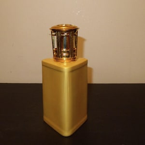 May include: A gold-coloured, square-shaped fragrance lamp with a decorative, gold-coloured top. The lamp has a geometric design and a shiny finish. The top has a cage-like design. The lamp is on a dark surface.