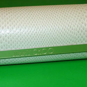 May include: A cylindrical, off-white case with a textured, snakeskin-like pattern. A green band with the brand name "JIMMY CHOO" is across the front. The case is on a bright green surface.