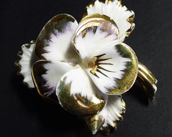 May include: A white porcelain flower brooch with gold accents. The flower has a delicate, hand-painted design with purple and white details. The brooch is a vintage piece and would make a beautiful addition to any jewelry collection.
