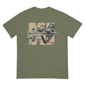 May include: A green t-shirt with a graphic of a tan and black submachine gun with a red dot sight. The graphic is surrounded by a tan and brown frame. The text "CAPACSKA ARMS" is below the graphic.