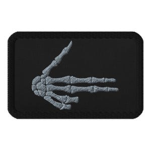 May include: A black rectangular patch featuring a gray embroidered skeleton hand giving a sideways gesture. The hand is detailed with visible bones and is set against a solid black background.