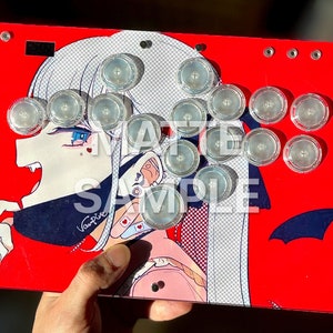 Peut inclure: Contr&ocirc;leur d'arcade rouge avec un graphisme de personnage d'anime vampire. Le contr&ocirc;leur a des boutons transparents et est &eacute;tiquet&eacute; "SAMPLE". Le design pr&eacute;sente un fond rouge avec un personnage blanc et bleu.