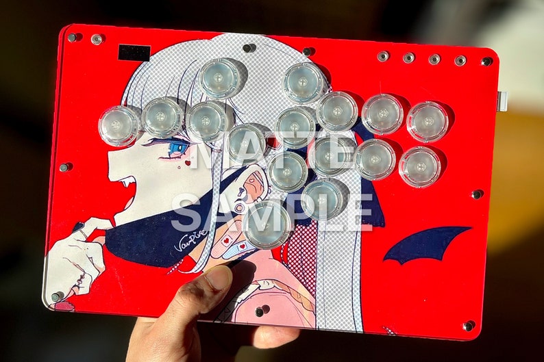 Peut inclure: Contr&ocirc;leur d'arcade rouge avec un graphisme de personnage d'anime vampire. Le contr&ocirc;leur a des boutons transparents et est &eacute;tiquet&eacute; "SAMPLE". Le design pr&eacute;sente un fond rouge avec un personnage blanc et bleu.