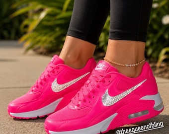 May include: A pair of bright neon pink Nike sneakers featuring a white midsole and a prominent Nike swoosh logo embellished with clear crystals. The text @theequeenofbling is visible in the lower right corner of the image.