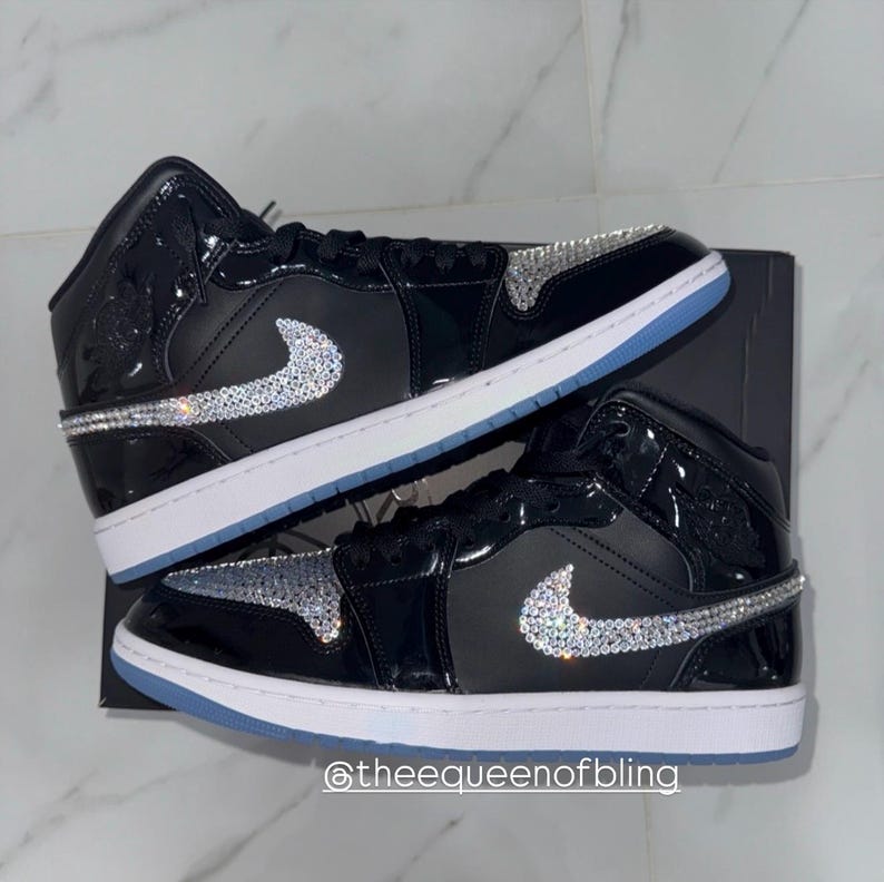 May include: A pair of black high-top sneakers featuring glossy patent leather panels and a white midsole. The side swoosh logos and the toe boxes are embellished with clear, reflective crystals. The text @theequeenofbling is visible at the bottom.