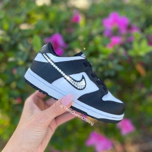 May include: Black and white low-top sneakers with a white swoosh decorated with clear rhinestones. The sneakers are on a hand with a white manicure.