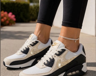May include: Nike Air Max 90 sneakers in white, beige, and black with gold-toned crystal embellishments on the side swoosh logos. The shoes feature white laces, a white midsole with a visible air unit, and black outsoles. Text reads @theequeenofbling.