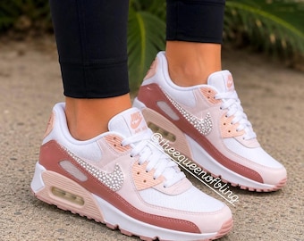 May include: Nike Air Max 90 sneakers in white, light pink, and dusty rose featuring a crystal-embellished Nike swoosh logo on the side. The shoes have white laces, a visible air unit in the heel, and the text theequeenofbling printed on the side.