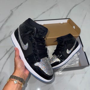May include: A pair of black high-top sneakers featuring a shiny patent leather finish, black laces, and a white sole. The toe box and the side swoosh logo are embellished with clear, sparkling crystals. The text @theequeenofbling is visible.