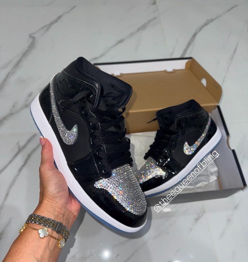 May include: A pair of black high-top sneakers featuring a shiny patent leather finish, black laces, and a white sole. The toe box and the side swoosh logo are embellished with clear, sparkling crystals. The text @theequeenofbling is visible.