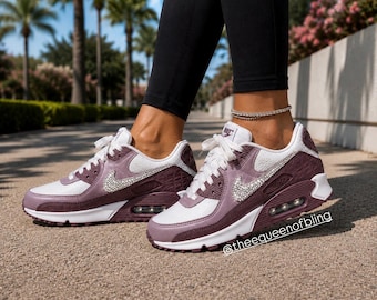 May include: A pair of mauve and white sneakers featuring a prominent Nike swoosh logo embellished with clear crystals on the side. The shoes have white laces, a visible air cushioning unit in the heel, and the text @theequeenofbling on the side.