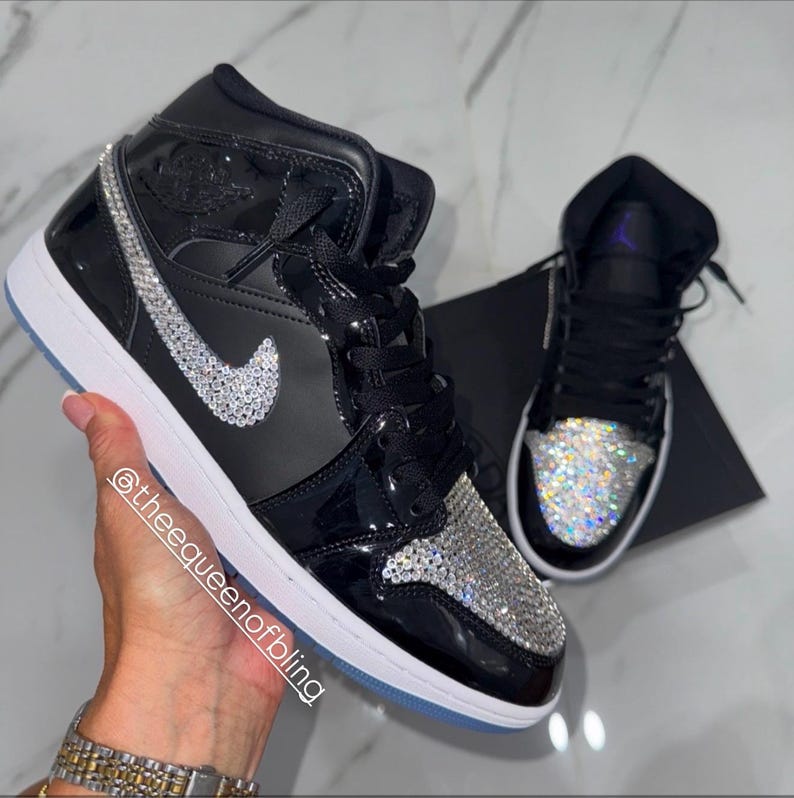 May include: A pair of black high-top sneakers featuring a glossy finish, white midsoles, and clear crystal embellishments covering the toe box and the side swoosh logo. The text @theequeenofbling is printed diagonally across the side of the shoe.