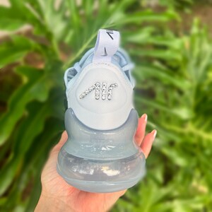 Puede incluir: Una zapatilla Nike Air Max 270 azul claro con suela de burbuja de aire transparente. La zapatilla tiene un logotipo Swoosh blanco en el lateral y un logotipo "AIR" blanco en la parte posterior. El logotipo "AIR" est&aacute; decorado con pedrer&iacute;a.