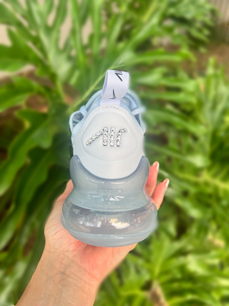 Puede incluir: Una zapatilla Nike Air Max 270 azul claro con suela de burbuja de aire transparente. La zapatilla tiene un logotipo Swoosh blanco en el lateral y un logotipo "AIR" blanco en la parte posterior. El logotipo "AIR" est&aacute; decorado con pedrer&iacute;a.