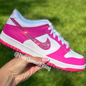 May include: White and pink low-top trainers with a pink swoosh decorated with pink rhinestones. The trainers have a white midsole and a pink outsole. The text "@TheeQueenOFBling" is visible on the side of the shoe.