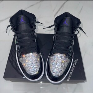May include: A pair of black high-top sneakers featuring a purple Jumpman logo on the tongue and a toe box covered in clear, reflective crystals. The sneakers have black laces and are displayed on a black box with the text @theequeenofbling.