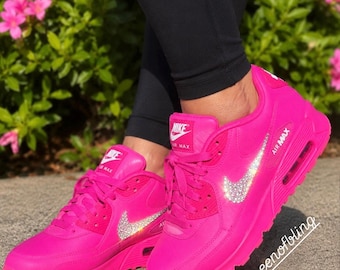 May include: A pair of bright hot pink Nike Air Max sneakers featuring a sparkling crystal-encrusted Nike swoosh logo on the side. The shoes have matching pink laces, a pink sole with a visible air unit, and the text @theequeenofbling on the image.