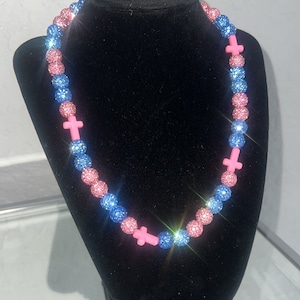 May include: A pink and blue beaded necklace with pink cross charms. The beads are sparkly and have a glittery finish.