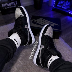 May include: A pair of black high-top sneakers featuring white soles, black laces, and silver rhinestone embellishments on the toe box and side swoosh logos. The sneakers are worn with white socks featuring a small black swoosh logo and black pants.