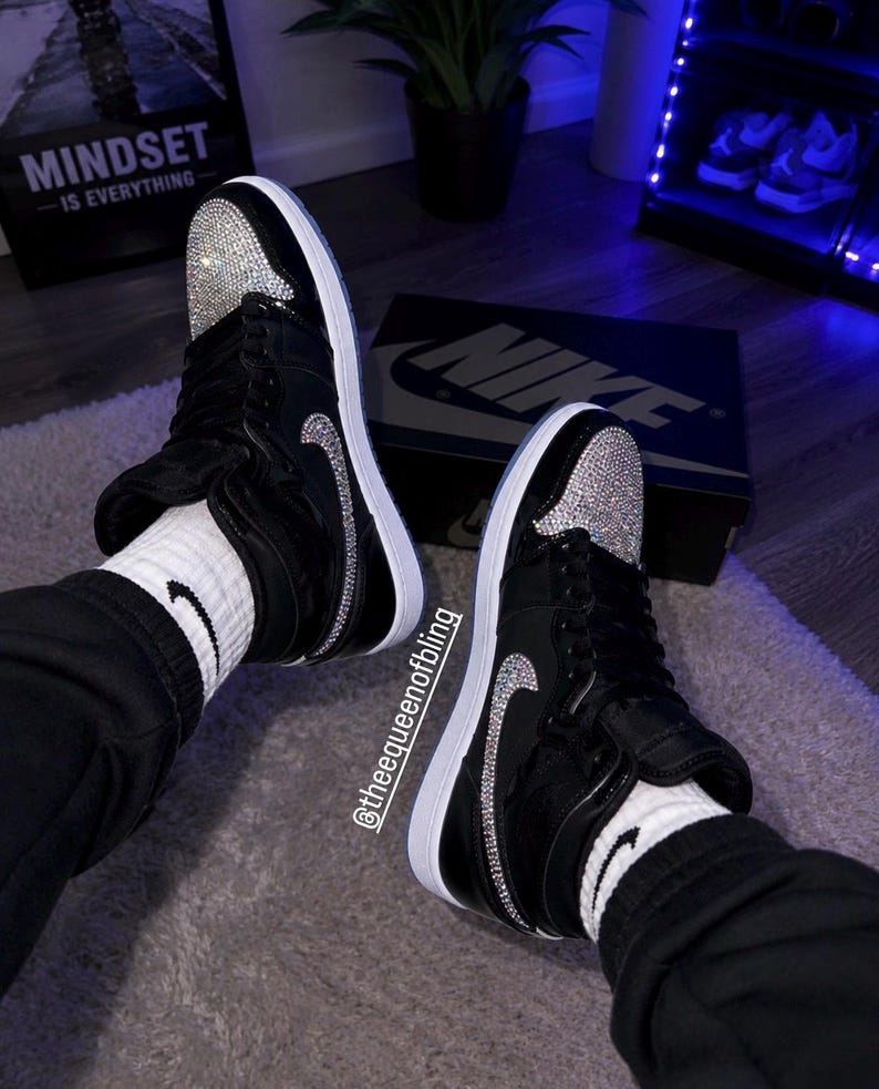 May include: A pair of black high-top sneakers featuring white soles, black laces, and silver rhinestone embellishments on the toe box and side swoosh logos. The sneakers are worn with white socks featuring a small black swoosh logo and black pants.