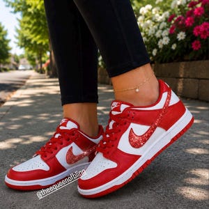 May include: A pair of red and white low-top sneakers featuring red glitter Nike swoosh logos on the sides, red laces, and white midsoles. The text @theequeenofbling is visible on the lower left side of the image near the front sneaker.