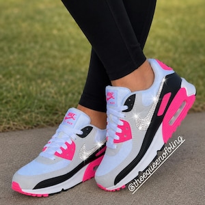 May include: A pair of Nike Air Max sneakers featuring a white, grey, and black color scheme with vibrant pink accents. The side Nike swoosh logo is embellished with clear crystals. The tongue features a Nike Max label, and the text @theequeenofbling is visible.