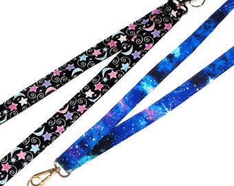 May include: Two fabric lanyards with metal clasps. One lanyard is black with pink and blue stars and crescent moons. The other lanyard is blue with a galaxy print.