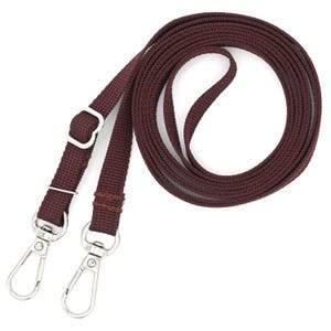 K&ouml;nnte beinhalten: Ein braunes gewebtes Band mit zwei silbernen Drehverschl&uuml;ssen. Das Band ist aufgerollt und bereit, an einer Tasche oder Handtasche befestigt zu werden.