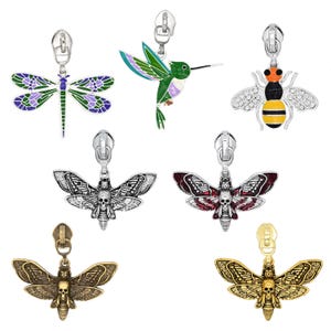 May include: A collection of seven metal zipper pulls featuring various designs including a purple and green dragonfly, a green hummingbird, a striped bee, and four moth-shaped pulls with skull details in silver, red, bronze, and gold tones.