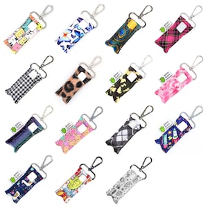 Könnte beinhalten: Eine Sammlung von Stoff-Lippenbalsamhaltern mit silbernen Karabinerhaken. Die Halterungen weisen verschiedene Muster auf, darunter Katzen, Schmetterlinge, Leopardenmuster, Karomuster und Weltraumdesigns. Jeder Halter hat ein kleines grünes Etikett mit Text.