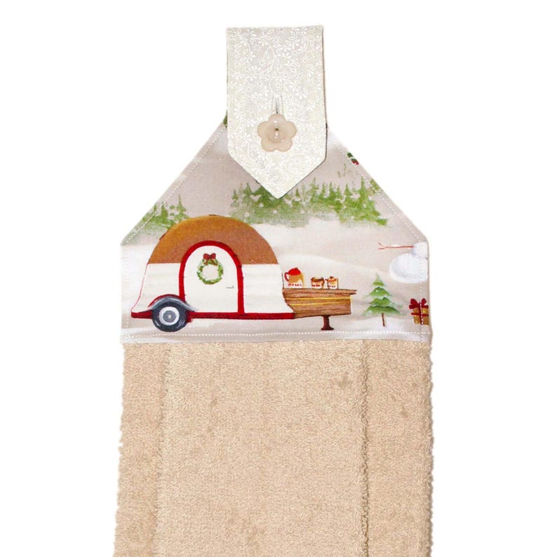 Puede incluir: Un pa&ntilde;o de cocina beige con una parte superior decorativa que representa una escena invernal con una caravana. La caravana es blanca y marr&oacute;n con ribete rojo, una corona en la puerta y una peque&ntilde;a mesa con golosinas. La parte superior tiene un bot&oacute;n floral blanco.