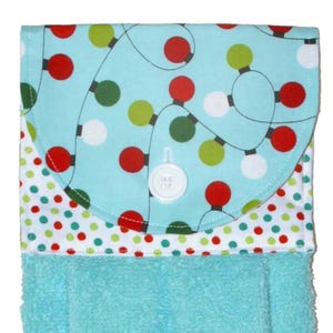 May include: A turquoise hand towel with a decorative flap featuring a pattern of red, green, and white Christmas lights on a light blue background. The flap is secured with a white button. The bottom of the towel is a solid turquoise color.
