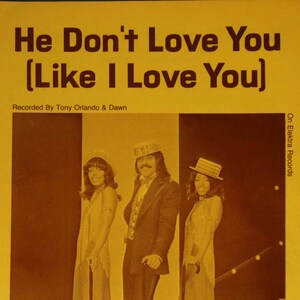May include: A vintage record album cover featuring a group of three people in a stage setting. The album cover is yellow with brown text that reads "He Don't Love You (Like I Love You)". The text "Recorded By Tony Orlando & Dawn" is also on the cover. The group is dressed in 1970s attire, with the man in the middle wearing a suit and a top hat. The women are wearing long dresses and hats. The album cover is from Elektra Records.