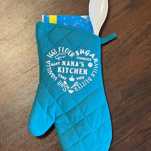 Może przedstawiać: Turkusowa, pikowana rękawica do piekarnika z białym tekstem, który brzmi "Nana's Kitchen", otoczona składnikami, takimi jak jajka, mąka, cukier, mleko, masło, sól, ciasto i ciasta.