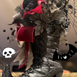 May include: A black lace-up boot with a high heel serving as a vase, filled with dark foliage, black roses, and a deep red velvet ribbon. The image features digital overlays of a spider web, a small spider, a white skull, and a black bat.