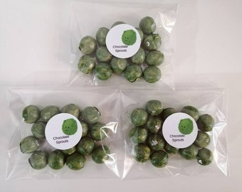 Può includere: Tre sacchetti di plastica trasparente pieni di caramelle verdi a forma di germoglio. Ogni sacchetto ha un adesivo rotondo bianco con un germoglio di cartone animato e le parole "Chocolate Sprouts".