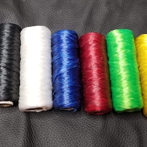 May include: Seven spools of waxed thread in various colors: purple, black, white, blue, red, green, and yellow. The spools are cylindrical and arranged side by side on a dark leather surface. Ideal for crafting and leatherwork.