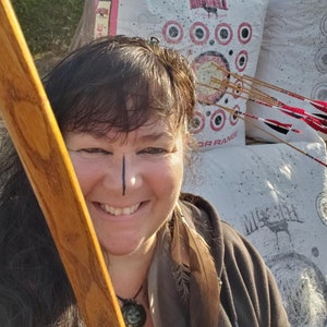 May include: A woman with dark hair and a black jacket is holding a wooden bow. She is smiling and looking at the camera. Behind her is a white archery target with red rings and several arrows sticking out of it. The target has the text "MORRELL" and "3D RANGE" printed on it.