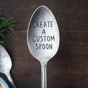 May include: A silver spoon with the text "CREATE A CUSTOM SPOON" in black. The spoon is in focus, with a second spoon and greenery in the background. A red ceramic bowl is visible in the upper right corner.