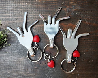 May include: Three silver fork keychains with red heart charms. The forks are bent into the shape of a hand with the fingers extended.