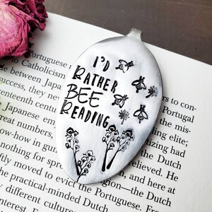 May include: A silver spoon bookmark with the text "I'd Rather Bee Reading" and a floral design. The spoon is on a book with text about Portuguese history.