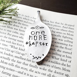 May include: A silver spoon with the words "One More Chapter" stamped on it. The spoon is a book mark and is resting on a page of a book.