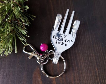 May include: A silver fork shaped into a hand with the words "MY FORKIN KEYS" stamped on it. The fork is attached to a silver key ring with a pink bead.