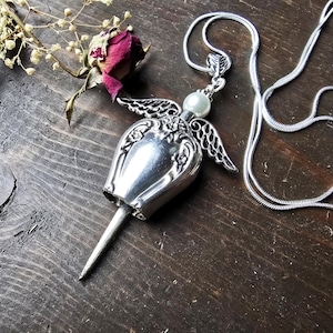 Pu&ograve; includere: Una collana con pendente in argento con un ciondolo a forma di campana, ali decorative e una perla. Il pendente &egrave; appeso a una delicata catena d'argento. Una rosa secca e rametti di fiori secchi sono accanto alla collana.