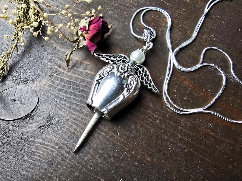Pu&ograve; includere: Una collana con pendente in argento con un ciondolo a forma di campana, ali decorative e una perla. Il pendente &egrave; appeso a una delicata catena d'argento. Una rosa secca e rametti di fiori secchi sono accanto alla collana.