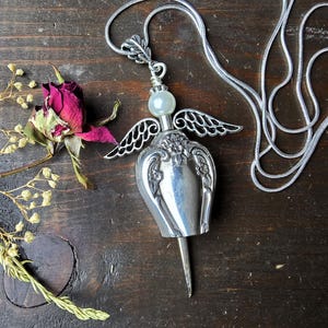 Pu&ograve; includere: Una collana con pendente a forma di angelo argentato con una perla madreperlacea e delicati dettagli alati. Il pendente &egrave; su una catena d'argento, su uno sfondo di legno scuro. Sono presenti anche una rosa secca e rametti di fiori secchi.