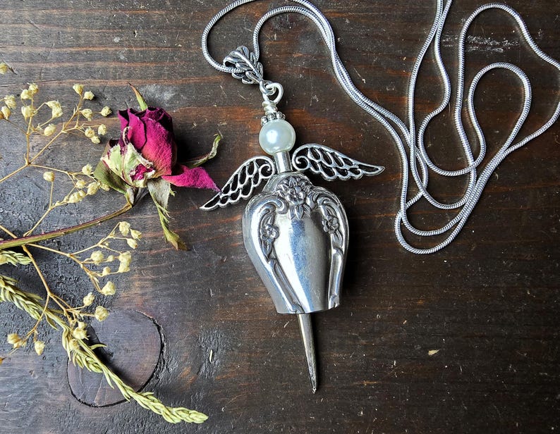 Pu&ograve; includere: Una collana con pendente a forma di angelo argentato con una perla madreperlacea e delicati dettagli alati. Il pendente &egrave; su una catena d'argento, su uno sfondo di legno scuro. Sono presenti anche una rosa secca e rametti di fiori secchi.