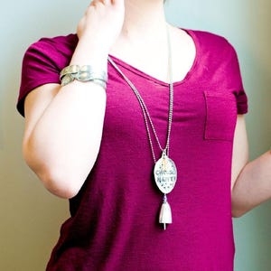 Pu&ograve; includere: Una maglietta magenta con tasca &egrave; abbinata a una collana e un braccialetto argentati. La collana presenta un ciondolo con le parole "CHOOSE HAPPY" e un ciondolo a campana. Il braccialetto ha un design strutturato unico.