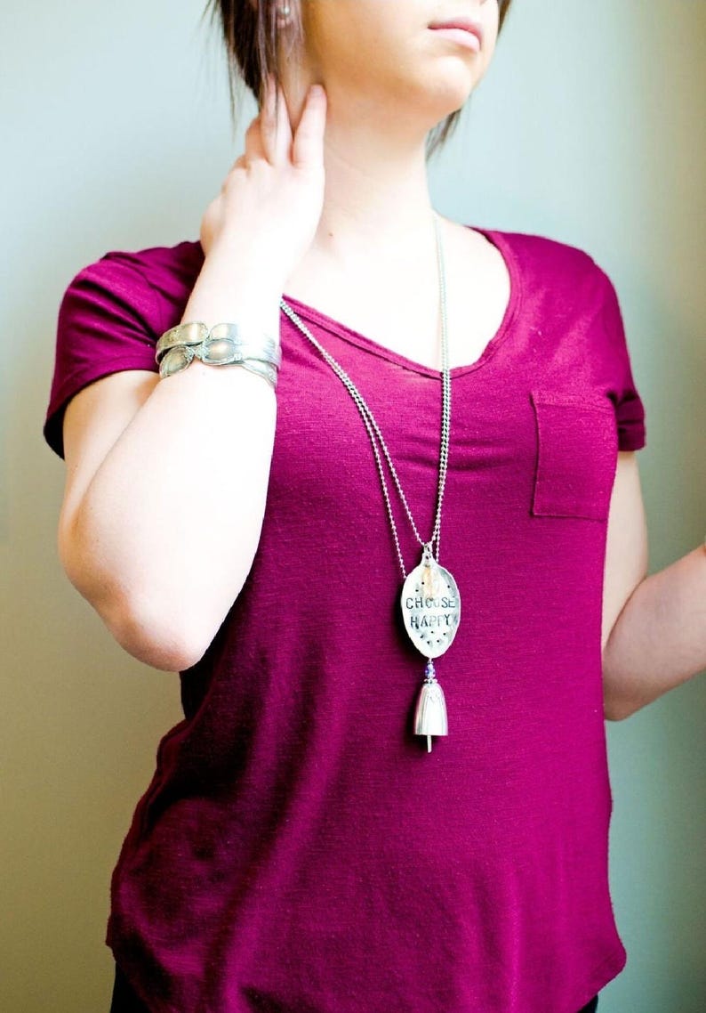Pu&ograve; includere: Una maglietta magenta con tasca &egrave; abbinata a una collana e un braccialetto argentati. La collana presenta un ciondolo con le parole "CHOOSE HAPPY" e un ciondolo a campana. Il braccialetto ha un design strutturato unico.